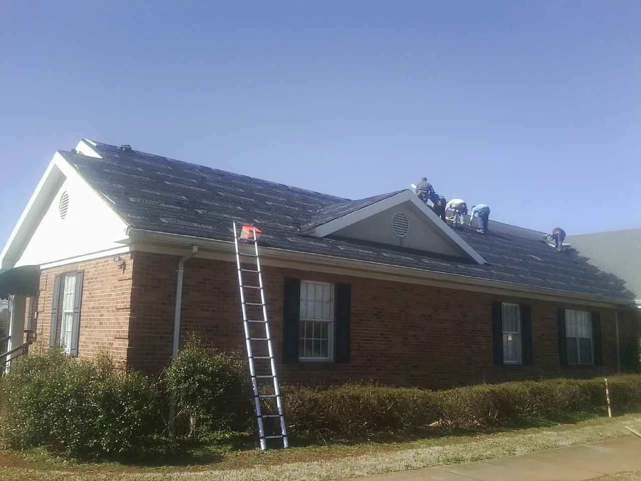 Crew tearing shingles off a brick ranch roof with a ladder set — Emergency Roof Repair work by our crew in La Motte