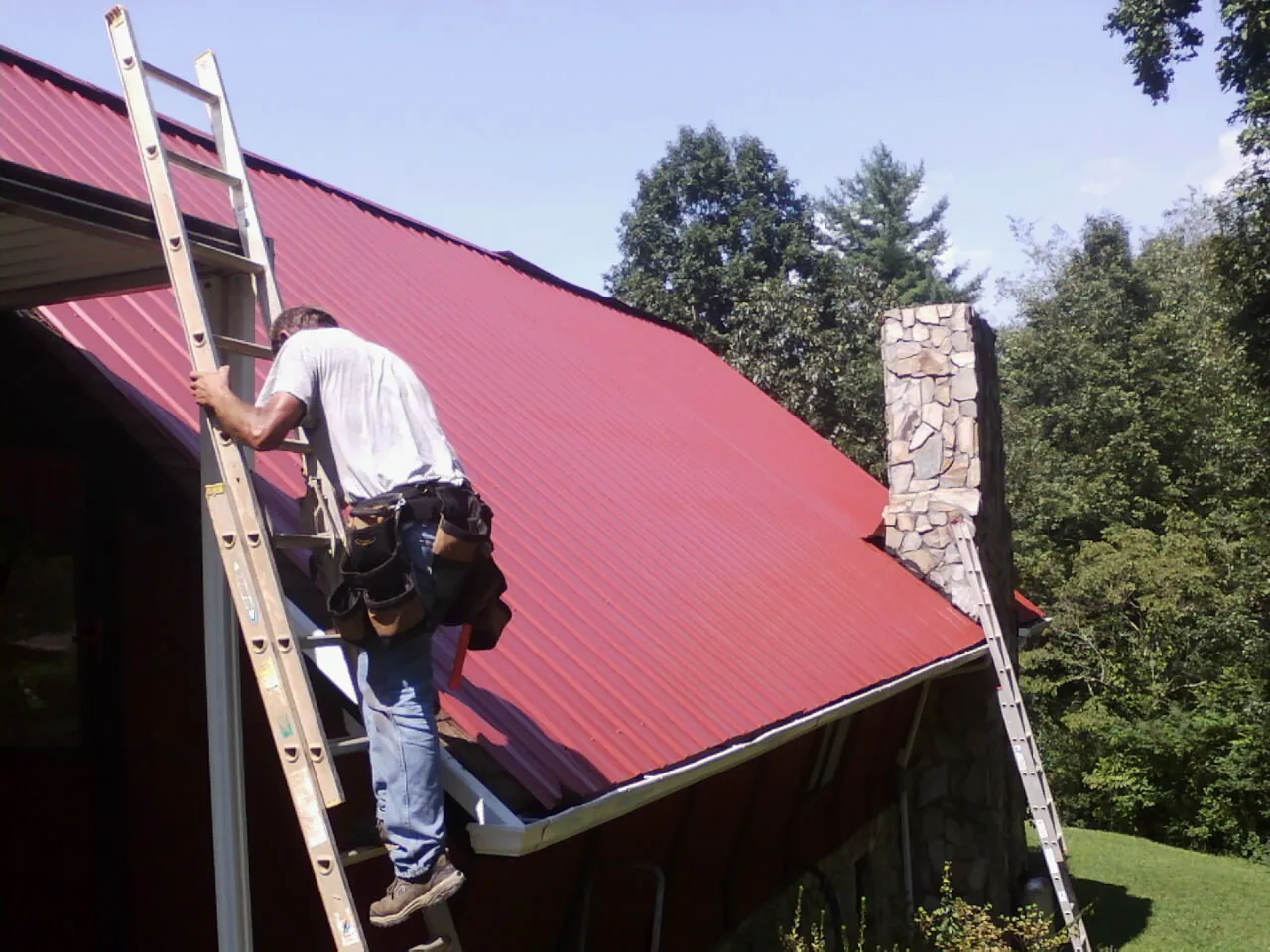 Roofer on a ladder beside a red metal roof and stone chimney — Soffit & Fascia Repair by Roofing Brothers in La Motte